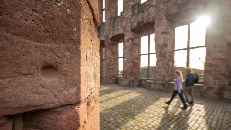 Een historische ruïne met grote ramen en zonlicht dat naar binnen valt. Twee personen lopen ontspannen over de stenen vloer.