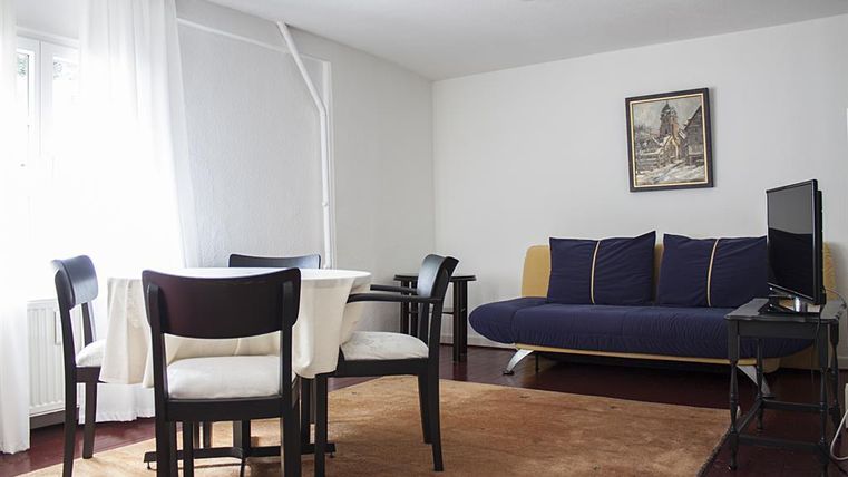 Een lichte woonkamer met een tafel en stoelen. Er is een bank en een televisie aan de wand.