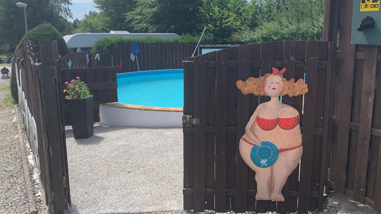 An entrance to a garden with a blue pool and a colorful mural of a happy woman in a red swimsuit. The area is well-maintained and has flowers in a pot.