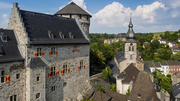 Eine alte Burg mit Steinfassade und bunten Bannern, umgeben von grünen Bäumen. Im Hintergrund ist ein Kirchturm und eine malerische Stadtlandschaft zu sehen.