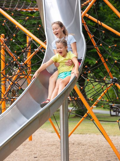 A mother and her child are happily sliding down a slide. In the background, colorful play equipment and a green park can be seen.