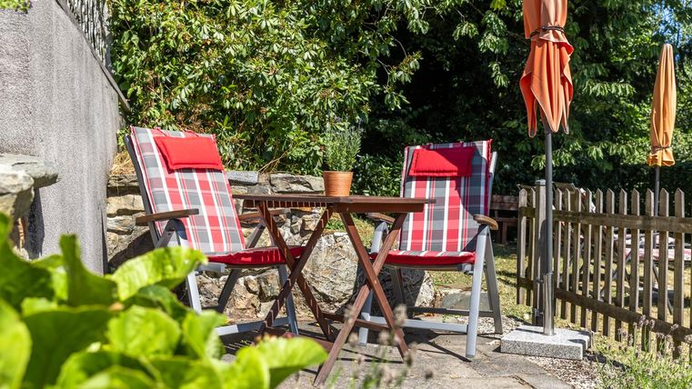 Een uitnodigende tuingedeelte met twee gestreepte stoelen en een kleine tafel. Op de achtergrond staan parasols en een houten schutting omlijst de ruimte.
