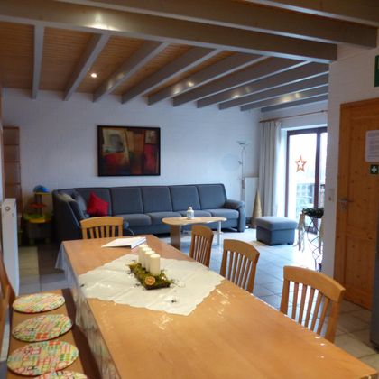 A modern kitchen with a large dining table and chairs. In the background, there is a cozy living room with a sofa and direct access to the balcony.