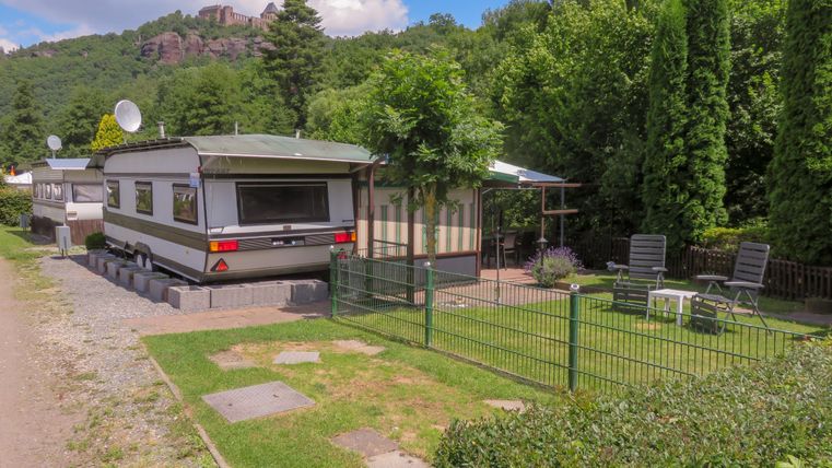 Ein Wohnwagen auf einem Campingplatz, umgeben von Bäumen und einem kleinen Garten. Es gibt zwei Liegen und einen Pavillon zum Entspannen.