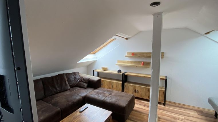 Ein moderner Dachzimmer mit einer bequemen Couch und einem Holz Tisch. Große Fenster sorgen für viel Tageslicht.