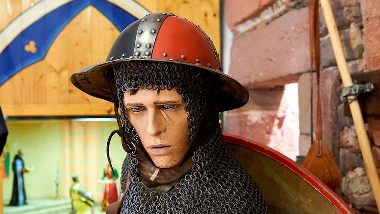 Een realistische figuur van een ridder met een kettingmail en een opvallende helm. Op de achtergrond zijn andere historische objecten zichtbaar.