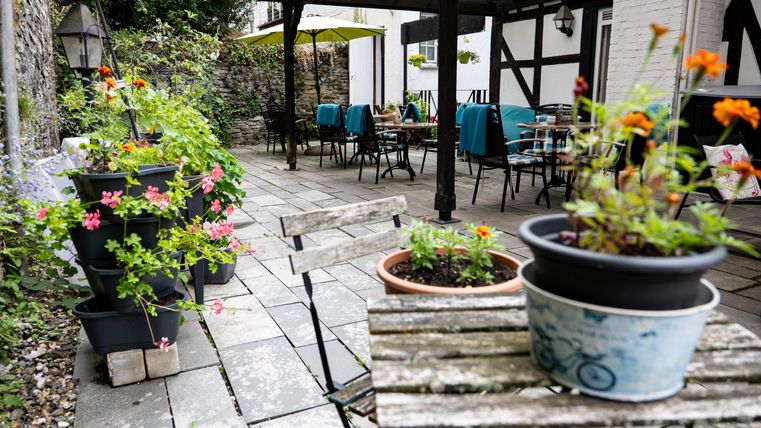 A cozy outdoor area with blooming plants and a paved floor. In the background, tables and chairs for guests can be seen.