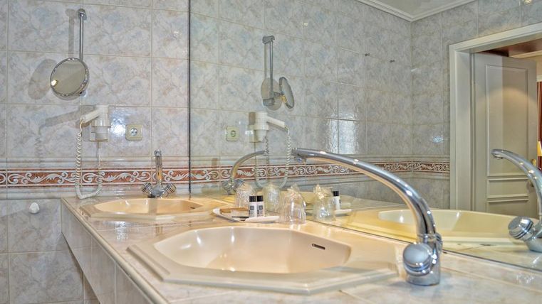 A modern bathroom with two sinks and a large mirror. The walls are lightly tiled and adorned with decorative elements.