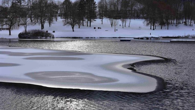 Een bevroren meer met een met sneeuw bedekte oever en bevroren water. Op de achtergrond zijn kale bomen en een rustige winterlandschap te zien.