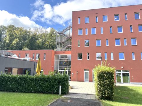 Ein modernes, orangefarbenes Gebäude steht in einer grünen Umgebung. Im Hintergrund sind Bäume und ein blauer Himmel sichtbar.