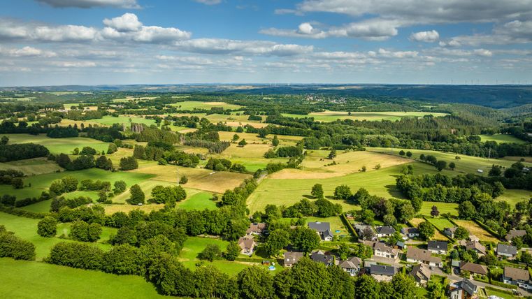 Eine weite Landschaft mit grünen Wiesen und sanften Hügeln. Im Vordergrund sind kleine Häuser und Bäume zu sehen.