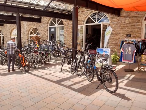 Een grote selectie fietsen staat in een groen binnenhof. Op de achtergrond zijn mensen en winkels zichtbaar.
