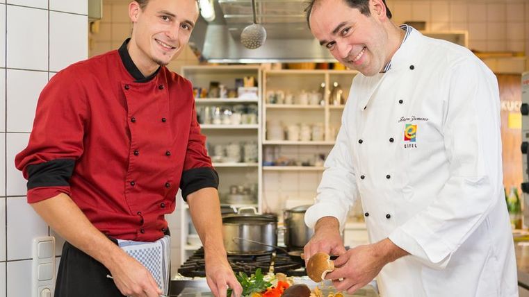 Two chefs are working in a modern kitchen. One is cutting fish while the other is preparing vegetables.