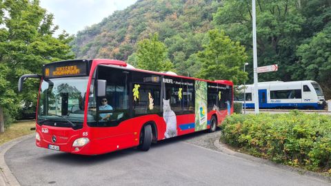 Een rode bus rijdt een bocht om terwijl een trein in de buurt stopt. Op de achtergrond zijn groene bomen en een bergachtige helling te zien.