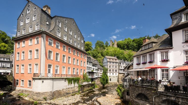 A picturesque town with historic buildings by the river. In the background, green hills and a blue sky can be seen.
