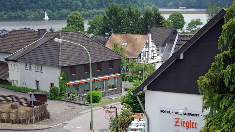 A picturesque view of a small village by the river. In the foreground, several houses and a restaurant can be seen, surrounded by green nature.