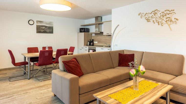Een moderne woonkamer met een beige bank en rode kussens. Op de achtergrond is er een open keuken met een eetgedeelte.