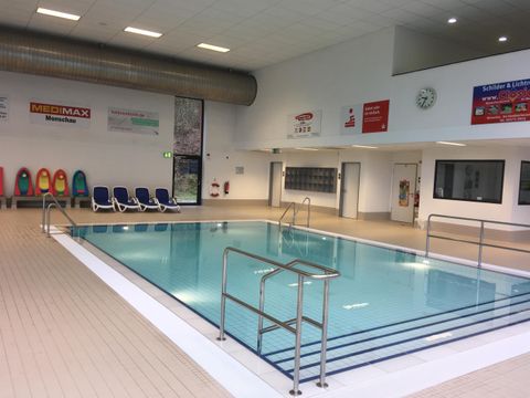 Ein helles Hallenbad mit klarem Wasser und einer Leiter. Es gibt Liegen am Rand des Schwimmbeckens.