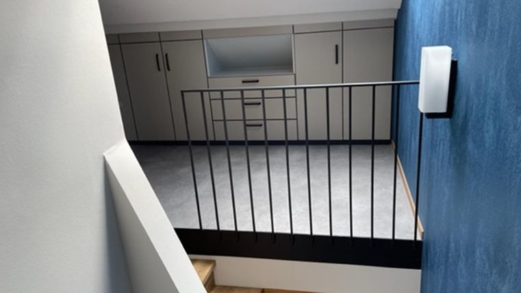 Een moderne ruimte met een blauwe muur en een houten trap. Aan de andere kant is er een lichte muur met kasten en een raam.