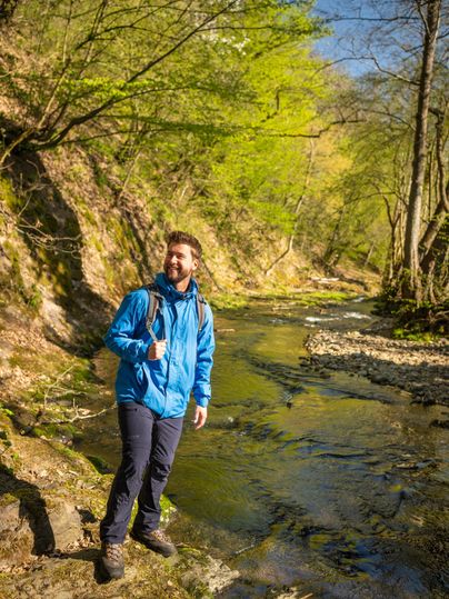Een man staat aan de oever van een heldere beek in een groene bos. Het is een zonnige dag en de omgeving voelt uitnodigend en vriendelijk aan.