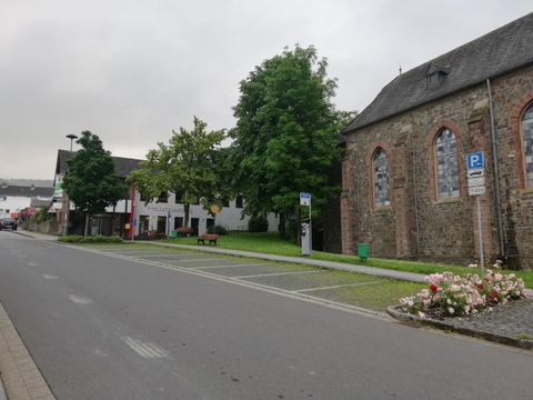 Eine ruhige Straße mit grünen Bäumen und einer alten Kirche. Links sind einige Gebäude und Blumenbeete zu sehen.