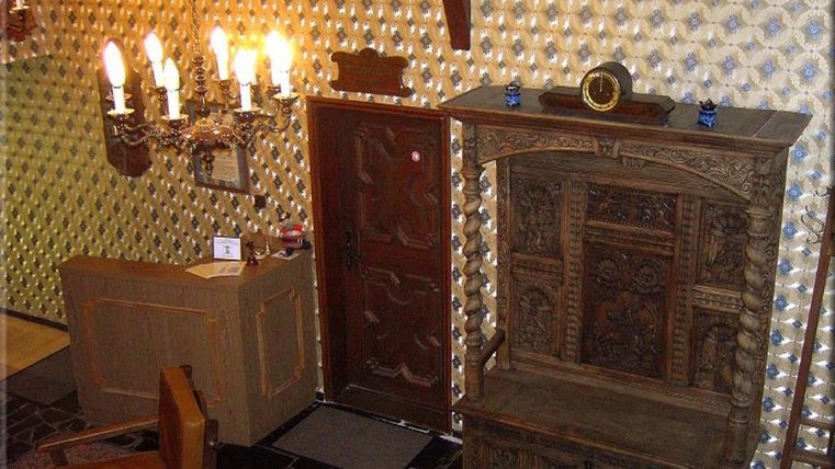 Ein gemütlicher Raum mit einer kunstvoll gestalteten Holzwand und einem traditionellen Schrank. An der Wand hängen Kerzenleuchter, und eine Eingangstür führt nach draußen.