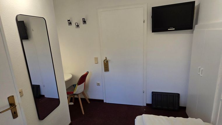 A simple hotel room with a bed, a table, and a television on the wall.
There is a large mirror and bright light from the ceiling.