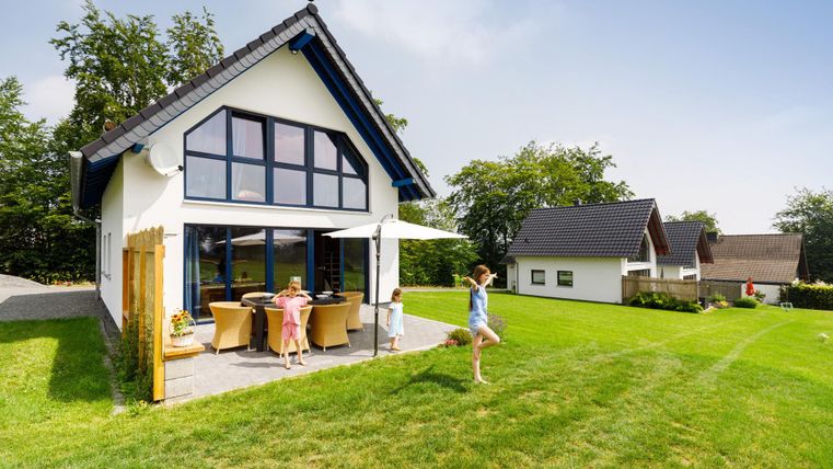 A modern house with large windows stands in a green garden. Children are playing on the lawn and enjoying the sun.