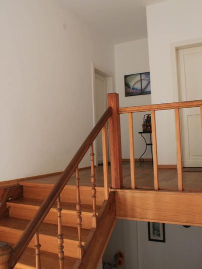 Een lichte gang met een houten trap en een leuning. Op de achtergrond zijn meerdere deuren en een tafel te zien.