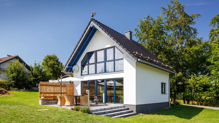 A modern house with large windows and a garden. The surroundings are green and sunny.