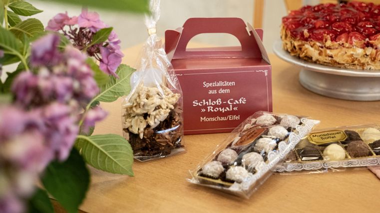 Ein Tisch mit verschiedenen Süßigkeiten und einer Torte. Im Vordergrund sind Pralinen und eine Papiertüte mit Knabbereien sichtbar.