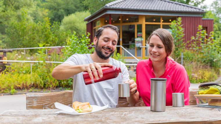 Ein glückliches Paar sitzt an einem Tisch und genießt ein Picknick in der Natur. Sie teilen sich Getränke und eine Mahlzeit in einer entspannten Umgebung.