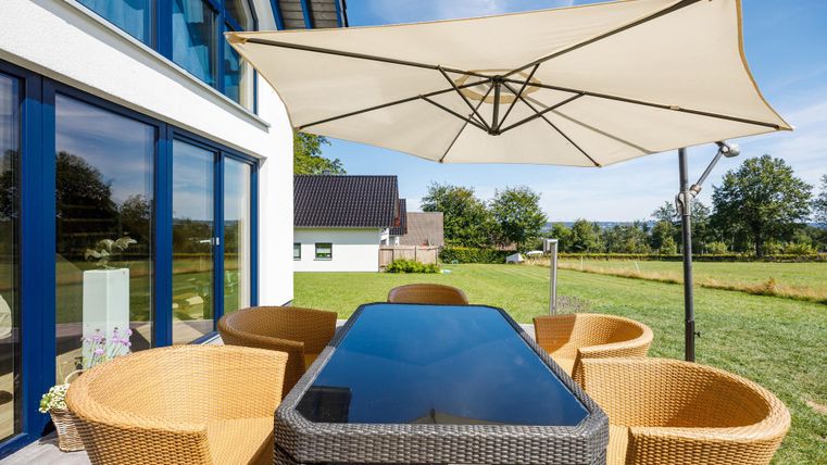 A beautiful outdoor dining table with wicker chairs and an umbrella. The surroundings are green and inviting.