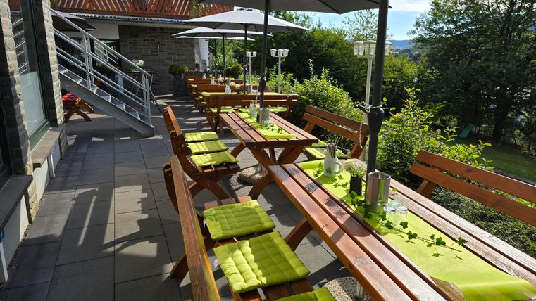 A cozy outdoor area with wooden tables and green umbrellas. The tables are decorated with colorful table runners and surrounded by bushes.