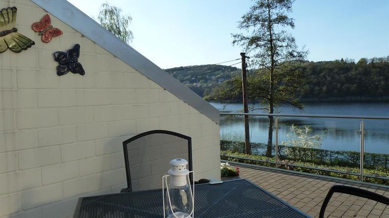 A cozy outdoor area with a table and a lantern. In the background, a calm river and green trees can be seen.