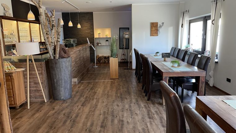 A modern restaurant with wooden flooring and large windows. Tables are set for guests and an inviting atmosphere is visible.