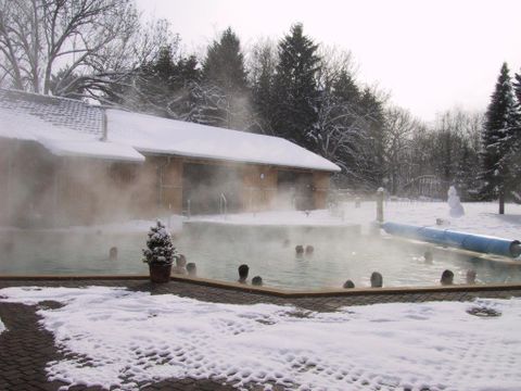 Ein beheiztes Schwimmbad im Freien, umgeben von Schnee und Dampf. Im Hintergrund steht ein Holzgebäude und ein paar Bäume sind sichtbar.