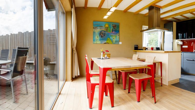 A bright kitchen with a dining table and red chairs. Through large windows, one looks out onto a beautiful garden.