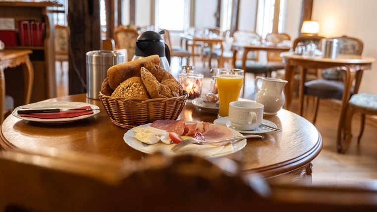 A laid table with fresh bread rolls, cold cuts, and a glass of orange juice. In the background, cozy chairs and a bright ambiance can be seen.