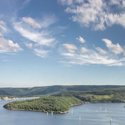 A picturesque landscape with a tranquil lake and gentle hills. Sailing boats glide across the water under a clear blue sky.