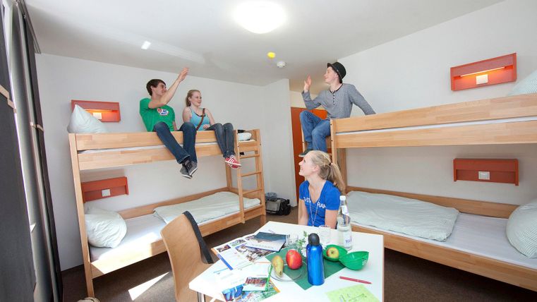 A lively youth hostel with two bunk beds and cheerful young people. They are playing and chatting, while snacks and drinks are on the table.