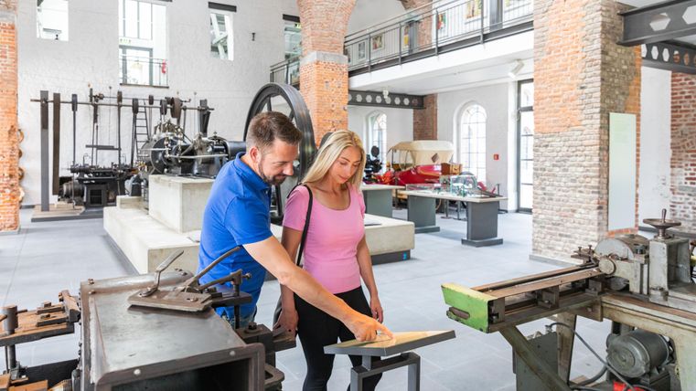 Twee personen bekijken samen een document in een museum voor oude machines. De ruimte is goed verlicht en toont historische apparaten.