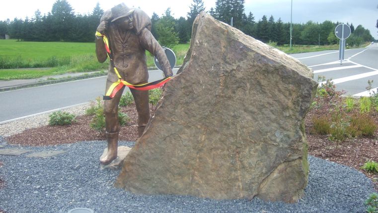 Een standbeeld van een arbeider die aan een grote steen werkt. De omgeving is groen met enkele struiken en een weg op de achtergrond.