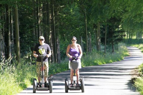 Two people are riding on Segways along a forest path. The surroundings are green and sunny.