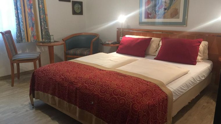 Een gezellige slaapkamer met een groot bed en rode kussens. Naast het bed staan een tafel en een stoel, evenals decoratieve schilderijen aan de muur.