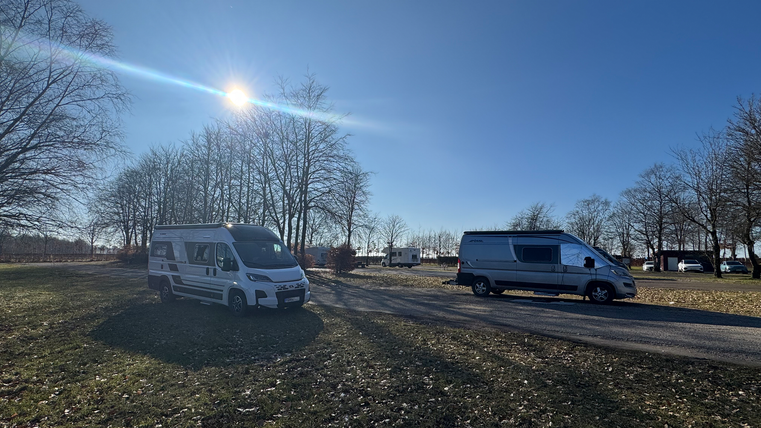 Twee campers staan op een plek met bomen op de achtergrond. De lucht is helder en zonnig.