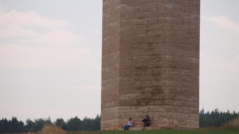 Een hoge, rechthoekige betonnen constructie staat op een groene heuvel. Twee personen zitten ervoor en genieten van het uitzicht.