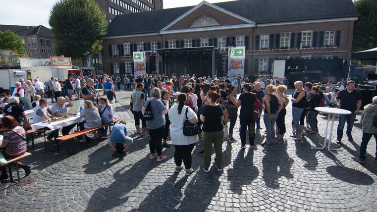 A large crowd gathers in a square in front of a building. In the background, stages and tables are set up, while the sun shines.