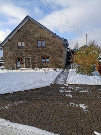 Ein charmantes Steinhaus umgeben von Schnee. Der Weg ist mit Pflastersteinen versehen und die Bäume sind winterlich karg.