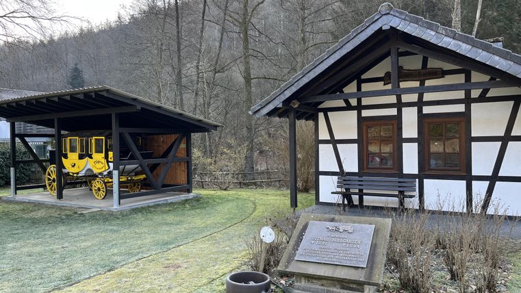 Ein traditionelles Fachwerkhaus steht neben einem überdachten Bereich mit einer historischen Kutsche. Die Umgebung ist grün und von Bäumen umgeben.